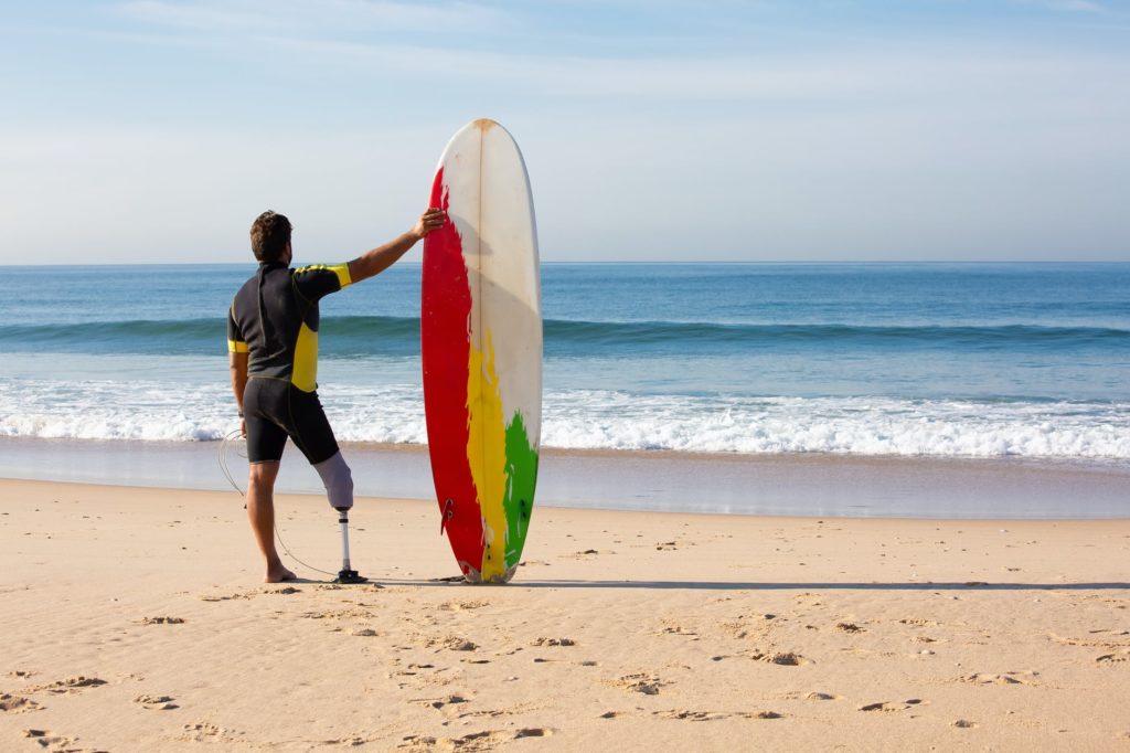 unrecognizable amputee man surfer with surfboard standing on seashore